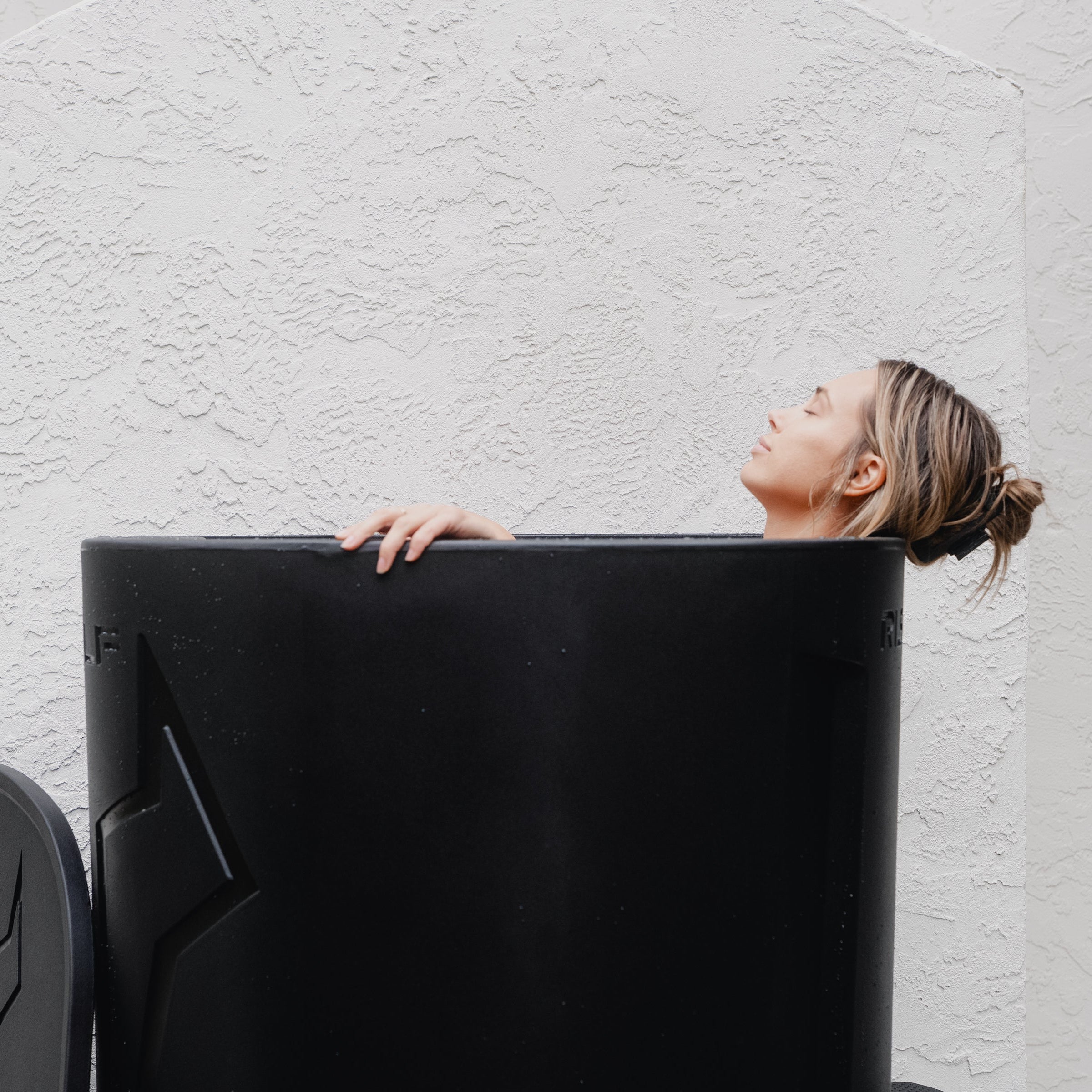 Woman lying inside a black cold plunge tub with head tilted back relaxing